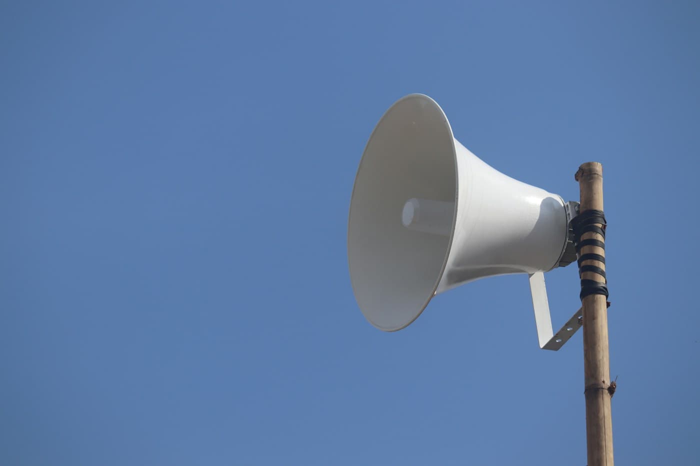 A white horn loudspeaker strapped to a wooden pole against a clear blue sky.
